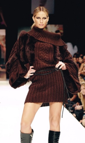 Modelo usando vestido de malha marrom e botas pretas em uma passarela de moda com uma multidão ao fundo.